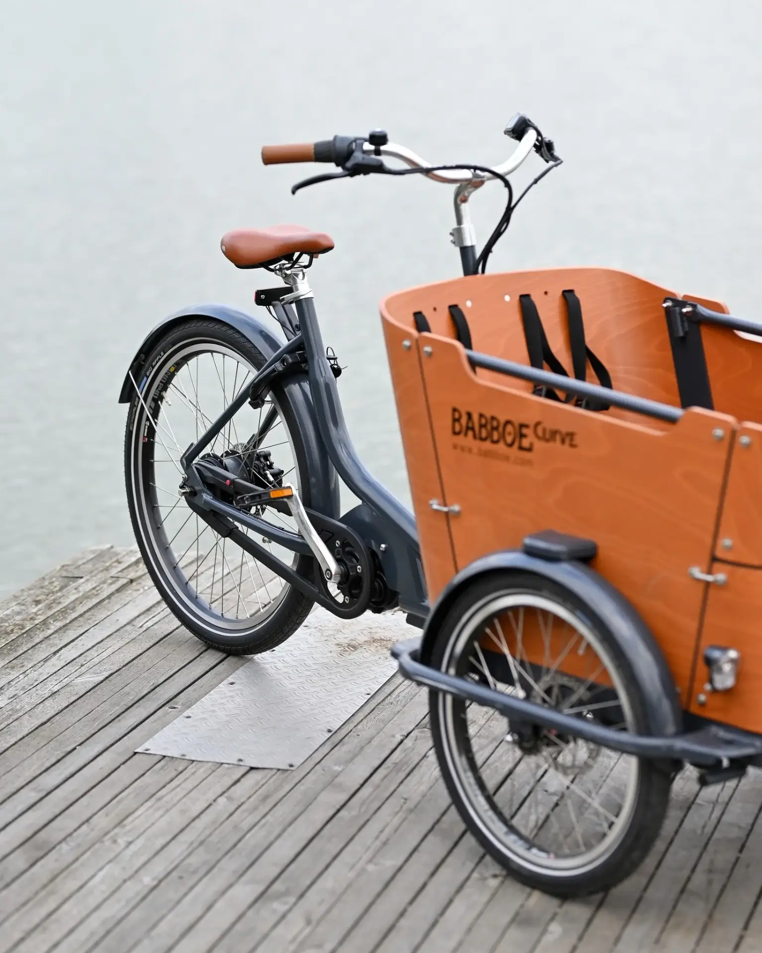 Kolmipyöräinen Babboe Curve -pyörä puulaatikolla seisoo puulaiturilla, jossa on harmaa runko, ruskea satula ja puinen laatikko.