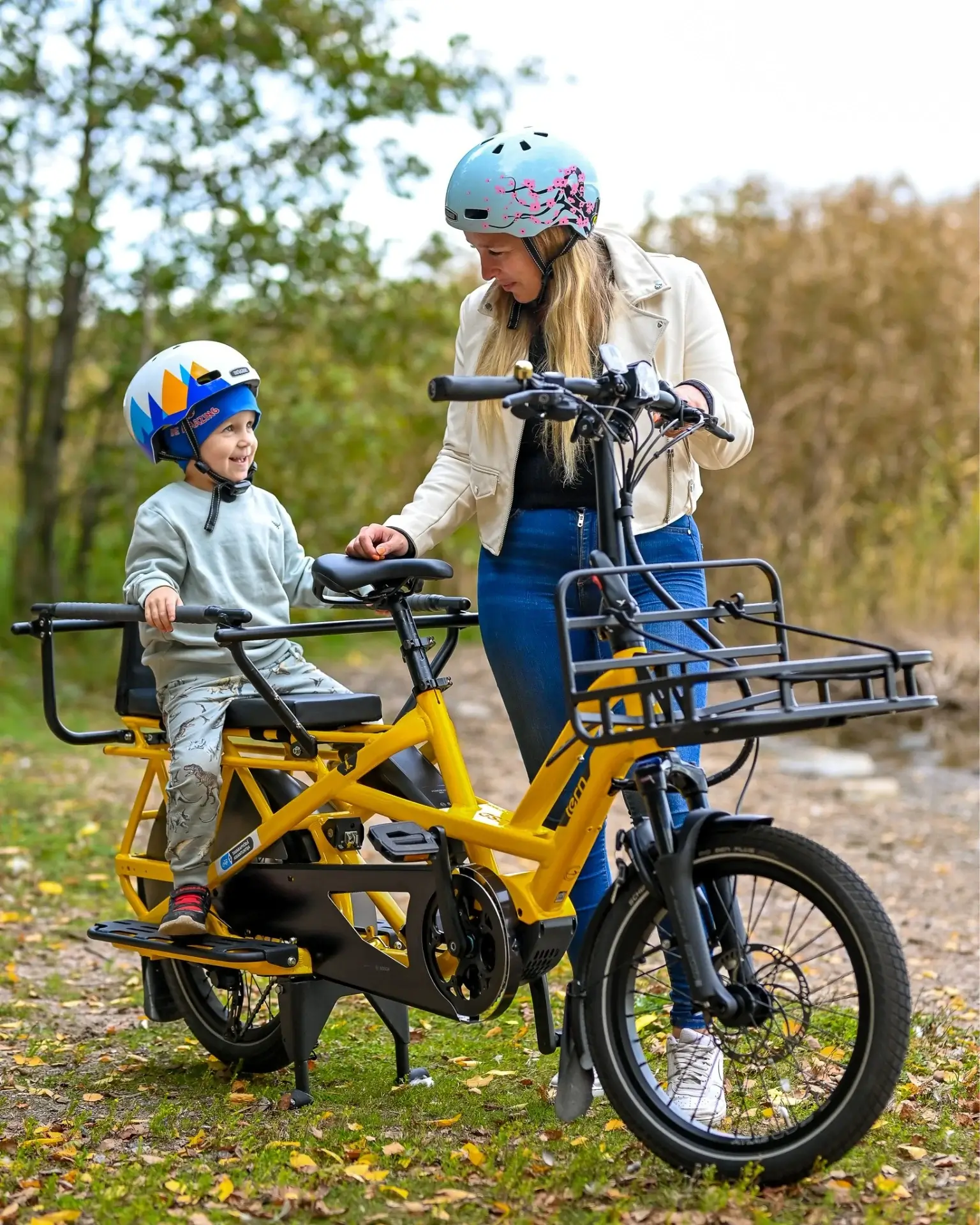 Äiti ja lapsi kypärät päässä keltaisella sähköpyörällä ulkona, aurinkoisessa säässä.
