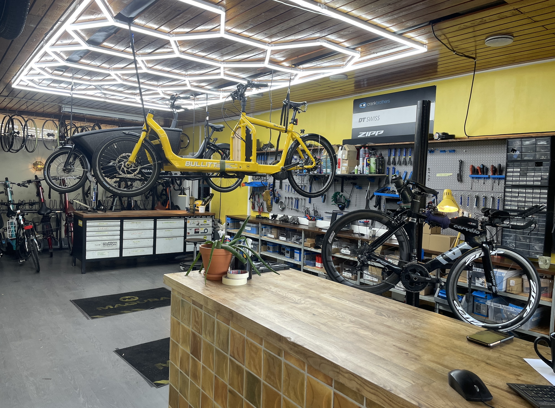 Polkupyöräliikkeen sisätiloissa on esillä polkupyöriä, työkaluja ja tarvikkeita. Kaksi polkupyörää on ripustettu katosta, ja työpöydällä on toinen polkupyörä. Tila on valaistu kirkkailla valoilla.