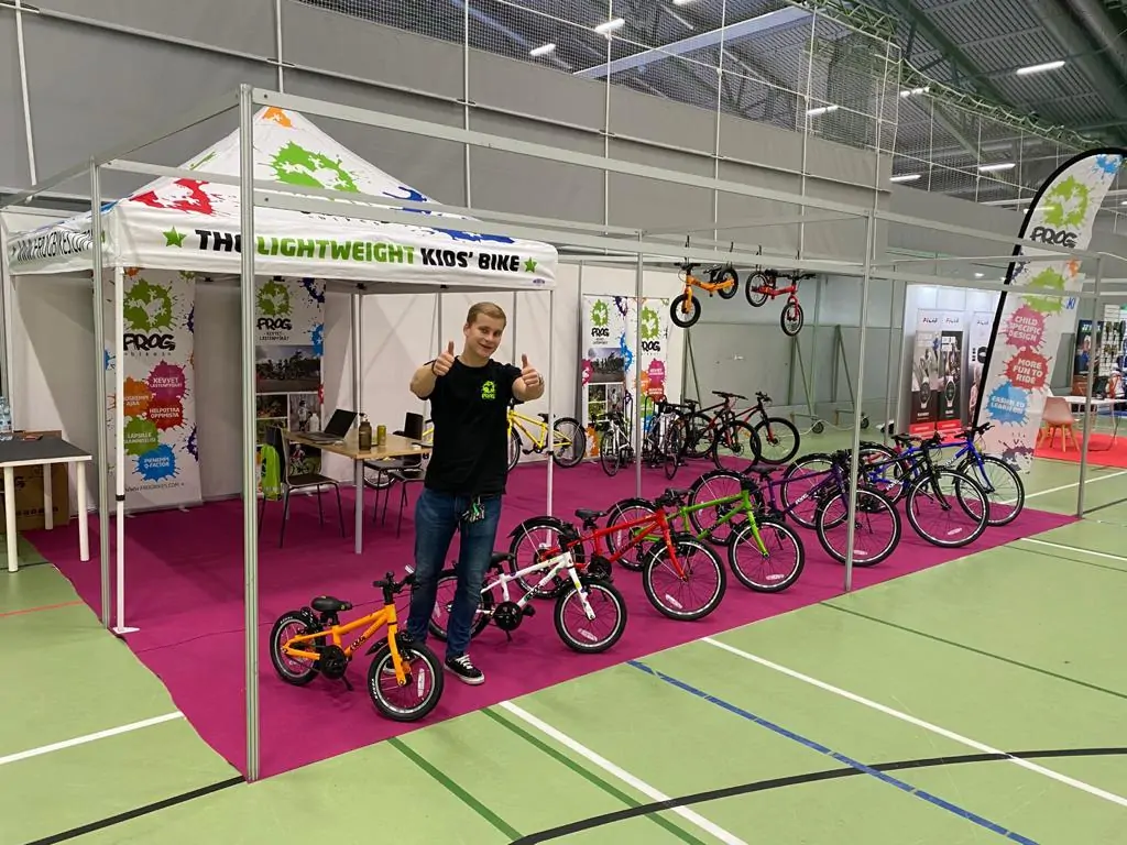 Messuhallissa on esillä lasten polkupyöriä, joita esittelee nuori mies peukut pystyssä. Pyörät ovat eri värisiä ja kokoisia, ja ne on aseteltu riviin vaaleanpunaiselle matolle. Taustalla näkyy messuosasto, jossa on pöytä ja tuoleja.