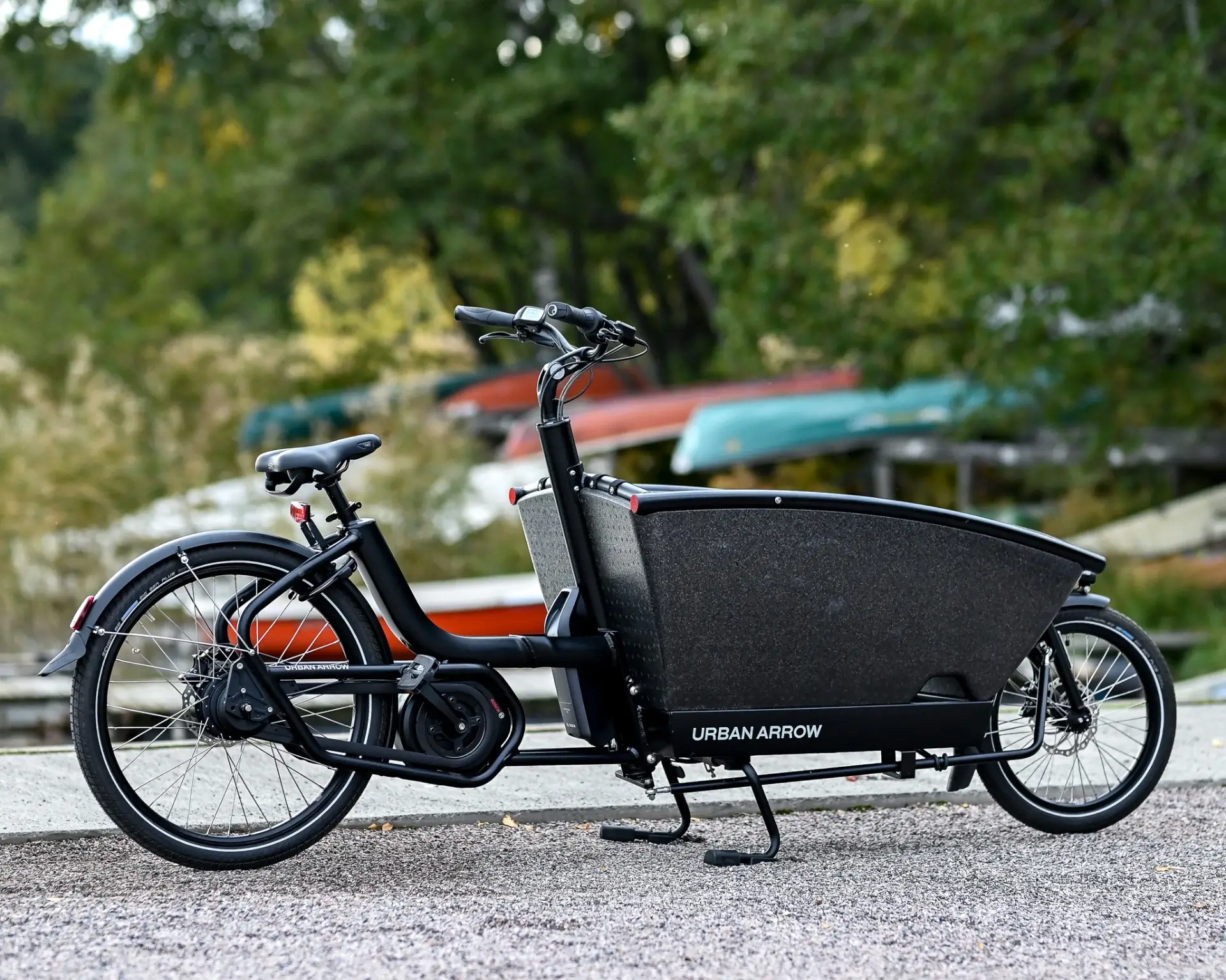 Musta Urban Arrow -sähköpyörä seisoo harmaalla alustalla, taustalla on puita ja veneitä.